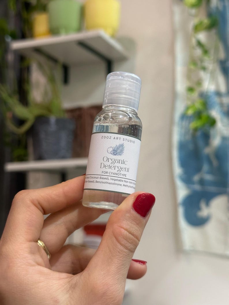 May include: A small, clear bottle of "Organic Detergent FOR CYANOTYPE" is held up. The label includes the text "ZOOZ ART STUDIO" and a botanical illustration. The bottle has a clear cap and contains a transparent liquid. The person holding the bottle has red nail polish.