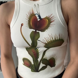 May include: White sleeveless tank top with a graphic print of a carnivorous plant with red and green details. The plant is depicted with a dragonfly on it.