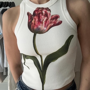 Könnte beinhalten: Ein weißes, geripptes Tanktop mit einem großen, detaillierten Grafikdruck einer einzelnen Tulpe in dunklen Rot-, Lila- und Rosatönen, mit einem langen grünen Stiel und Blättern, die sich von der Brustmitte bis zum Saum des Kleidungsstücks erstrecken.