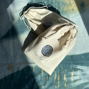 May include: Two beige drawstring bags with a spiral design in black ink. The bags are made of a natural fabric and have a simple, minimalist aesthetic. The text "Cyanotype A" is printed on one of the bags.