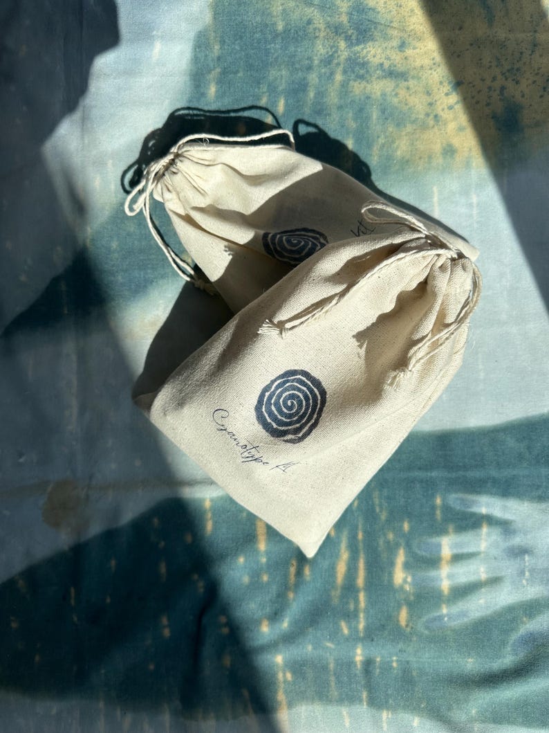 May include: Two beige drawstring bags with a spiral design in black ink. The bags are made of a natural fabric and have a simple, minimalist aesthetic. The text "Cyanotype A" is printed on one of the bags.