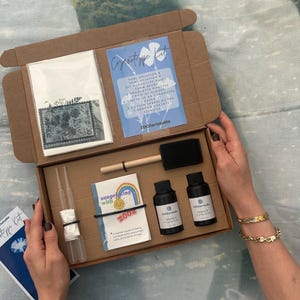 May include: An open cardboard box containing a cyanotype kit. The kit includes bottles of solution, a brush, pipettes, gloves, paper, fabric pieces, stencils, and a guide. The box is held open by a person.