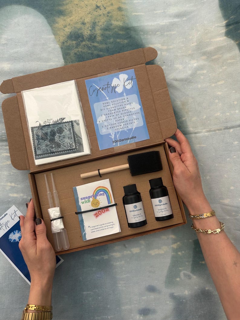 May include: An open cardboard box containing a cyanotype kit. The kit includes bottles of solution, a brush, pipettes, gloves, paper, fabric pieces, stencils, and a guide. The box is held open by a person.
