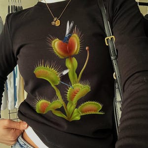 Op de afbeelding: Zwart shirt met lange mouwen met een kleurrijke afbeelding van een Venus vliegenval met een libel. De plant heeft groene, rode en gele details. Het shirt wordt gedragen met een spijkerbroek en een gouden ketting.