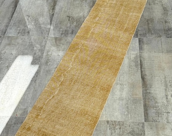 Puede incluir: Una alfombra de pasillo rectangular de color amarillo dorado con un aspecto envejecido y vintage. La alfombra tiene una superficie descolorida y texturizada y un borde blanco en los extremos cortos. La alfombra está colocada sobre un suelo de baldosas grises.