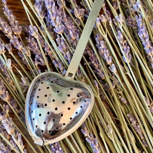 May include: A heart-shaped, silver tea infuser with a long handle and a heart-shaped cutout at the top. The infuser is perforated with small holes. The background features dried lavender sprigs.