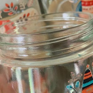 May include: A clear glass jar with a threaded neck for a lid. The exterior features a colorful graphic print of stylized trees, leaves, and floral elements in shades of orange, teal, and black, with small white accents throughout the design.