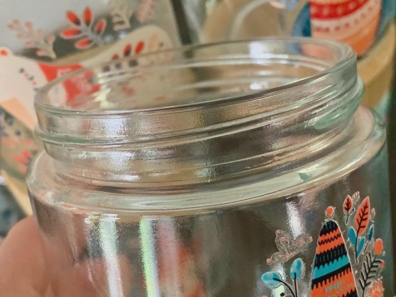 May include: A clear glass jar with a threaded neck for a lid. The exterior features a colorful graphic print of stylized trees, leaves, and floral elements in shades of orange, teal, and black, with small white accents throughout the design.