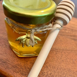 May include: A small glass jar filled with honey, with a gold lid and a bee charm. A wooden honey dipper is next to the jar.