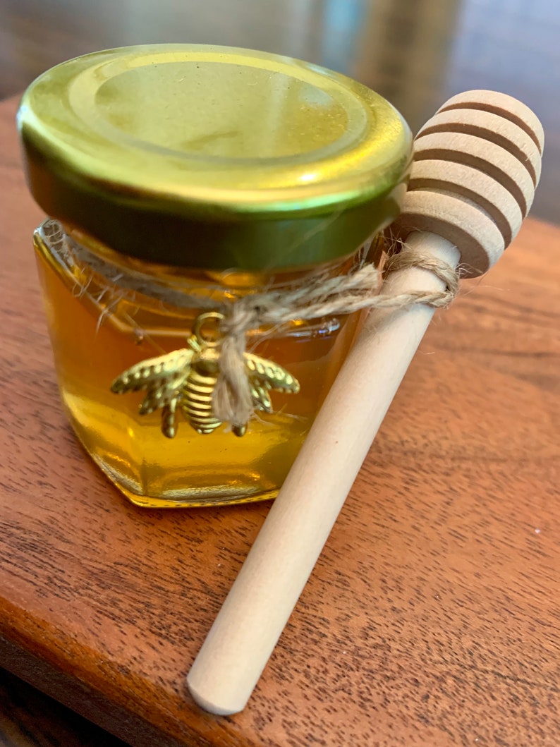 May include: A small glass jar filled with honey, with a gold lid and a bee charm. A wooden honey dipper is next to the jar.