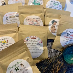 May include: A collection of small, tan, resealable pouches containing spice blends, each with a circular white label. Visible labels include Moroccan Blend, Adobo Blend, Taco Blend, Greek Blend, and Organic Thai Blend, featuring ingredient lists and gluten-free icons.