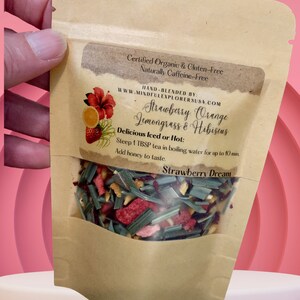 May include: A brown paper pouch with a clear window showing a blend of dried fruit, herbs, and flowers. The pouch is labelled "Strawberry Orange Lemongrass & Hibiscus" and "Strawberry Dream". It is certified organic, gluten-free, and naturally caffeine-free.