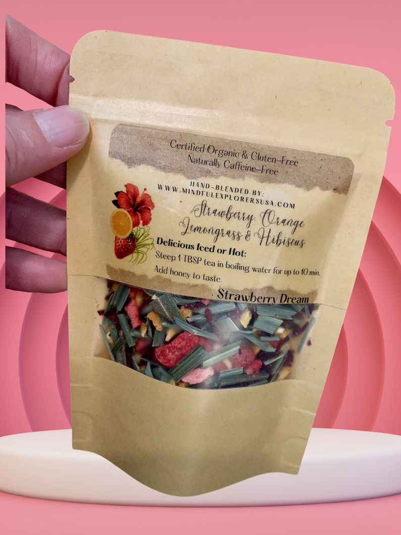 May include: A brown paper pouch with a clear window showing a blend of dried fruit, herbs, and flowers. The pouch is labelled "Strawberry Orange Lemongrass & Hibiscus" and "Strawberry Dream". It is certified organic, gluten-free, and naturally caffeine-free.