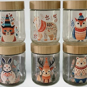 May include: Six clear glass jars with light brown wooden lids, arranged in two rows of three. Each jar features a colorful illustration of a woodland animal, including a cat, bear, owl, rabbit, deer, and another bear, all decorated with floral patterns.