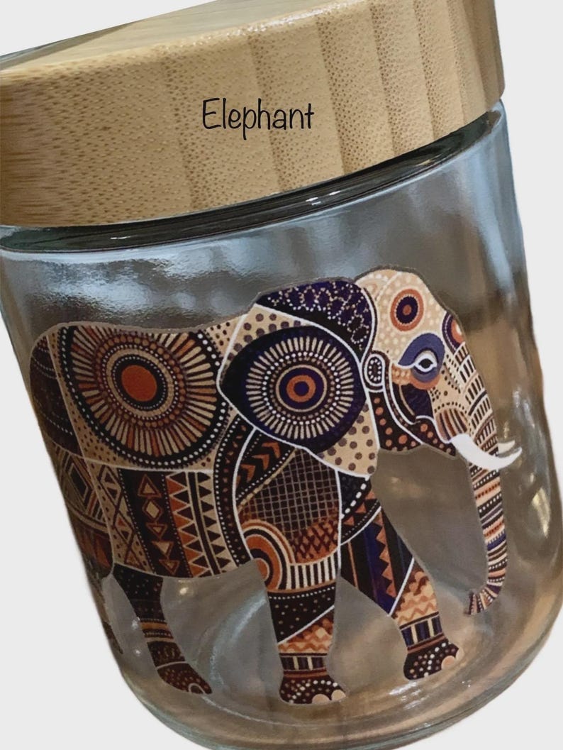 May include: A clear glass storage jar with a light brown bamboo lid. The word Elephant is printed on the lid. The side of the jar features a detailed graphic of an elephant decorated with intricate geometric patterns in shades of brown, orange, and black.