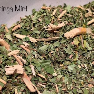 May include: Close-up of a blend of dried moringa leaves and mint, with a light brown color and a slightly rough texture. The text "Moringa Mint" is visible in the top left corner of the image.