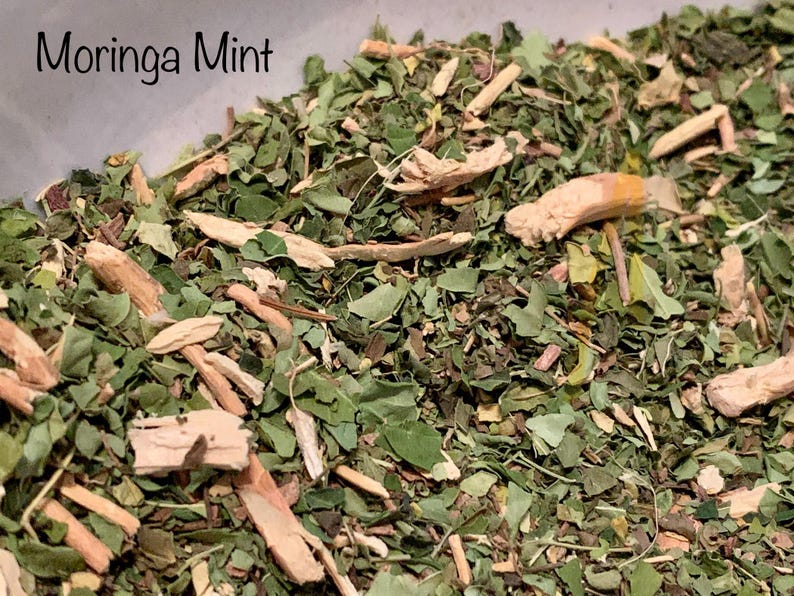 May include: Close-up of a blend of dried moringa leaves and mint, with a light brown color and a slightly rough texture. The text "Moringa Mint" is visible in the top left corner of the image.