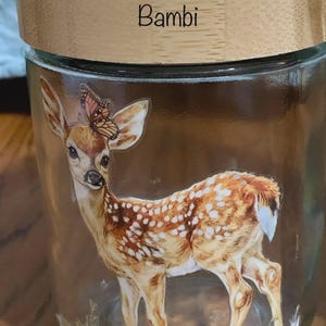 May include: A clear glass jar with a light-colored wooden lid featuring the name Bambi printed in black. The jar displays a detailed illustration of a spotted fawn with a monarch butterfly resting on its ear, set against a grassy base.