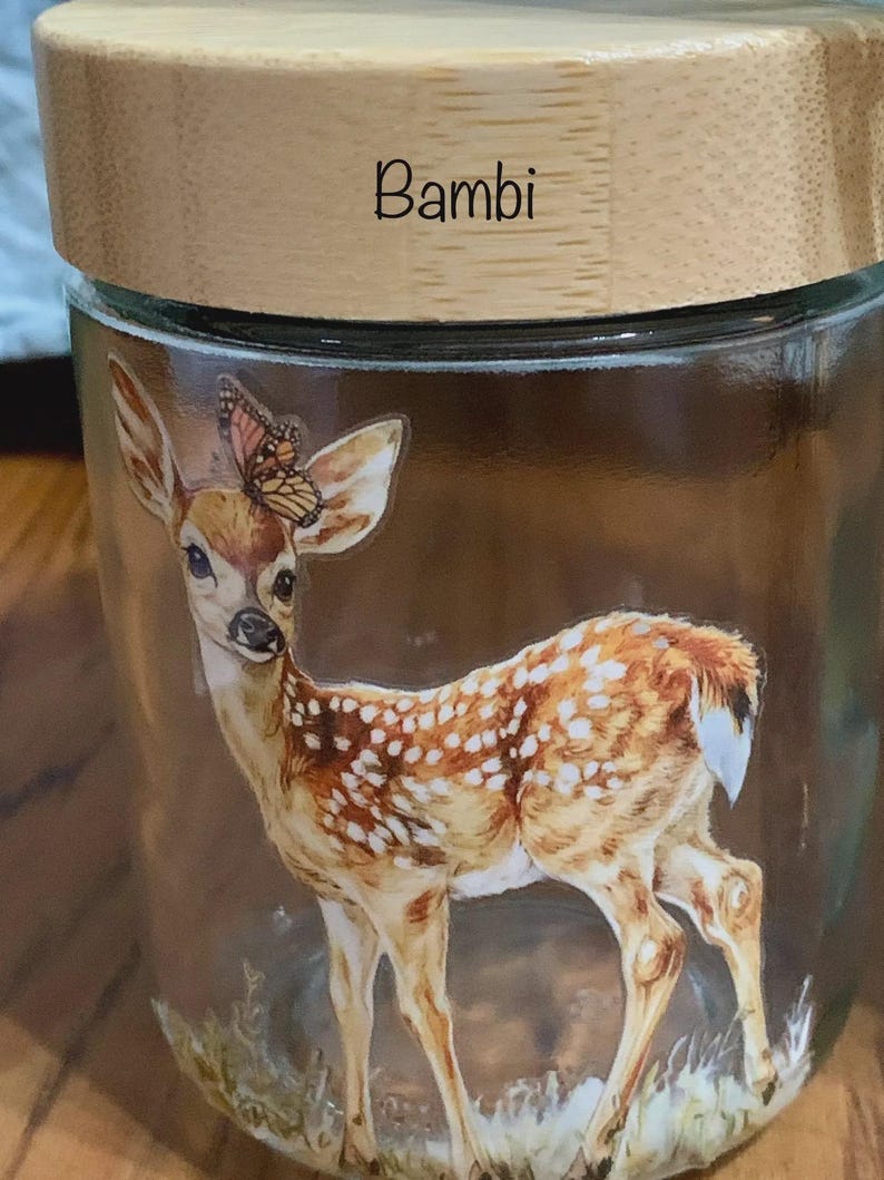May include: A clear glass jar with a light-colored wooden lid featuring the name Bambi printed in black. The jar displays a detailed illustration of a spotted fawn with a monarch butterfly resting on its ear, set against a grassy base.