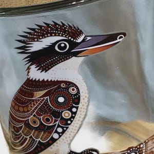 May include: A clear glass container featuring a detailed illustration of a kookaburra bird with brown, white, and black patterned plumage. The word Kookaburra is printed in black text above the bird, which is perched on a decorative branch.