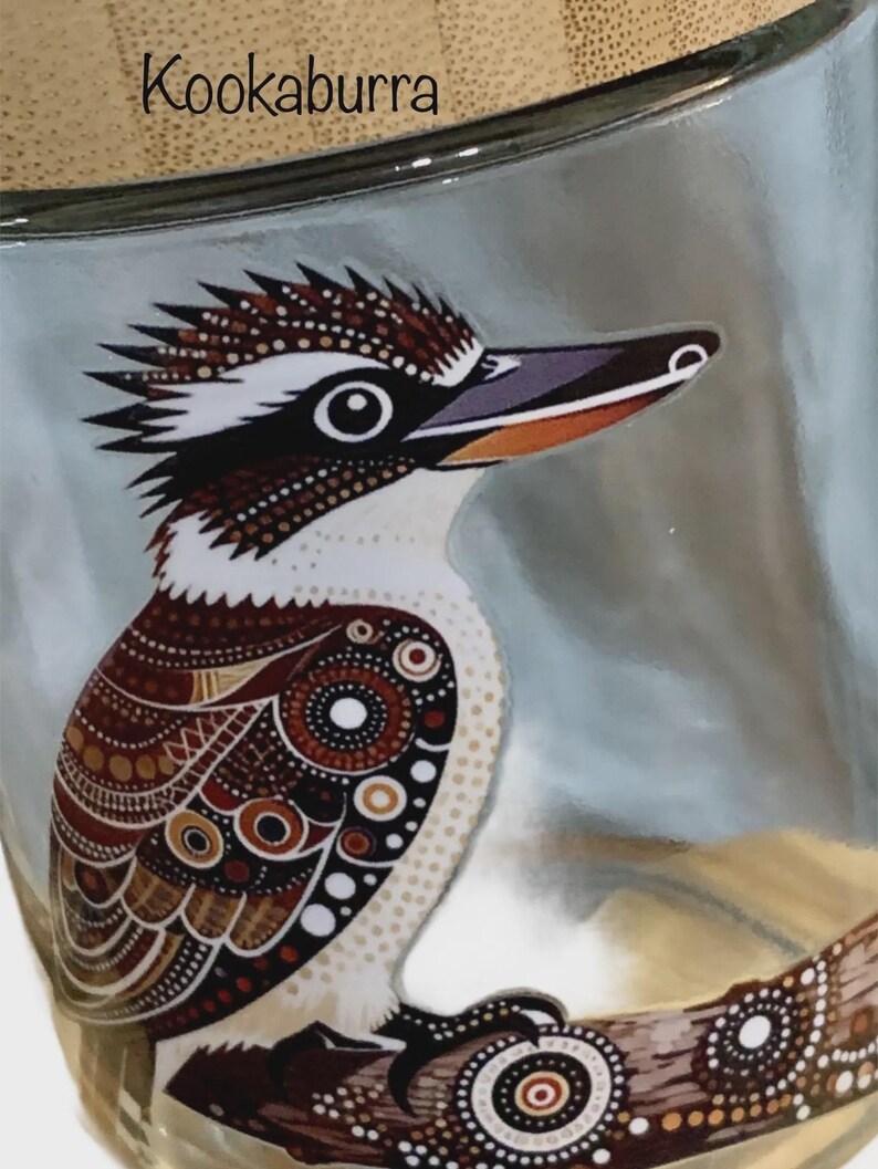 May include: A clear glass container featuring a detailed illustration of a kookaburra bird with brown, white, and black patterned plumage. The word Kookaburra is printed in black text above the bird, which is perched on a decorative branch.