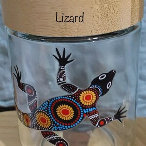May include: A clear glass storage jar with a light-colored wooden lid. The word Lizard is printed in black on the lid. The glass features a colorful lizard graphic decorated with intricate dot patterns in red, yellow, blue, and white colors.