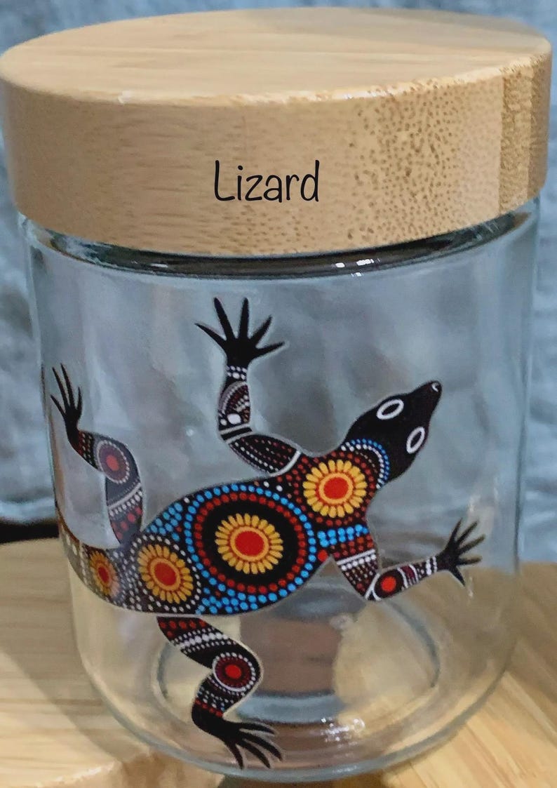 May include: A clear glass storage jar with a light-colored wooden lid. The word Lizard is printed in black on the lid. The glass features a colorful lizard graphic decorated with intricate dot patterns in red, yellow, blue, and white colors.