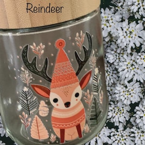 May include: A clear glass jar with a light-colored wooden lid featuring the word Reindeer printed on it. The jar displays a colorful graphic of a reindeer wearing a patterned hat, surrounded by illustrations of various trees and foliage.
