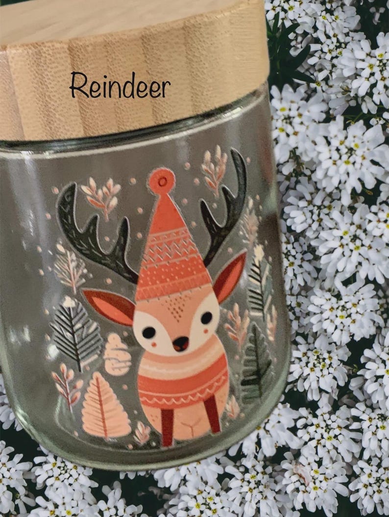 May include: A clear glass jar with a light-colored wooden lid featuring the word Reindeer printed on it. The jar displays a colorful graphic of a reindeer wearing a patterned hat, surrounded by illustrations of various trees and foliage.