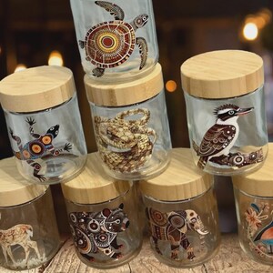 May include: A collection of seven clear glass storage jars with light-colored wooden lids. Each jar features a unique animal illustration, including a sea turtle, lizard, snake, kookaburra, deer, bear, and elephant, decorated with intricate patterns.