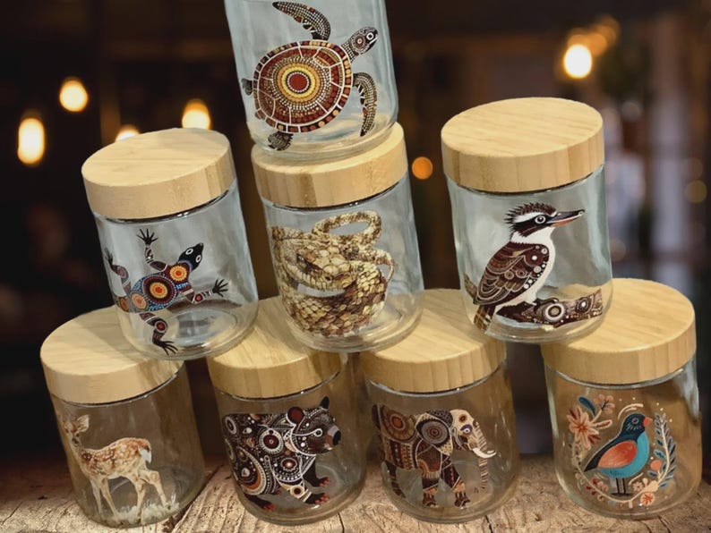 May include: A collection of seven clear glass storage jars with light-colored wooden lids. Each jar features a unique animal illustration, including a sea turtle, lizard, snake, kookaburra, deer, bear, and elephant, decorated with intricate patterns.