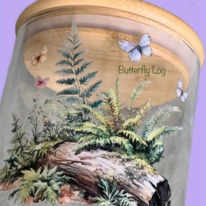 May include: A clear glass storage jar with a light-colored wooden lid. The exterior features a printed design of a fallen log covered in green ferns and various plants, accented by several butterflies and the text Butterfly Log in green.