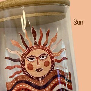 May include: A clear glass storage jar with a light-colored wooden lid. The jar features a graphic decal of a stylized sun with a human face, surrounded by wavy rays and layered, patterned bands in shades of brown, cream, and dark red. The word Sun is printed in brown text to the right of the lid.