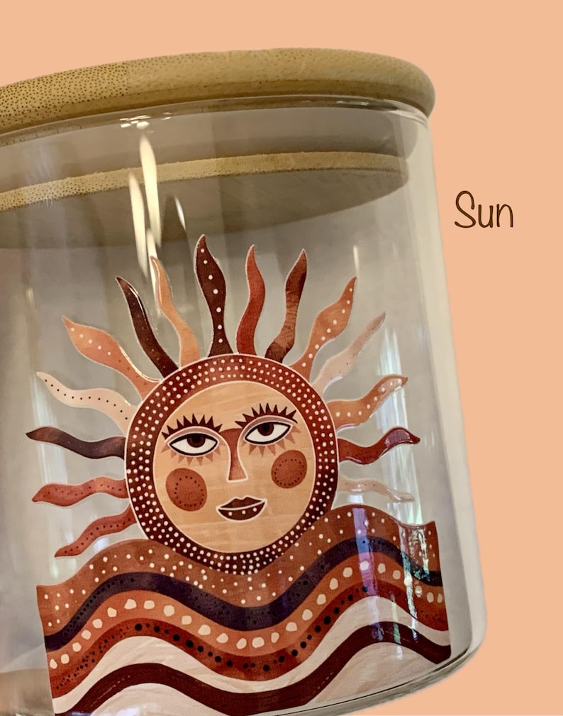 May include: A clear glass storage jar with a light-colored wooden lid. The jar features a graphic decal of a stylized sun with a human face, surrounded by wavy rays and layered, patterned bands in shades of brown, cream, and dark red. The word Sun is printed in brown text to the right of the lid.