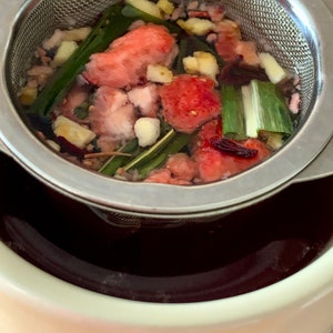 May include: A stainless steel tea infuser filled with a mixture of red and green ingredients, including fruit, herbs, and spices. The infuser is sitting in a white ceramic mug.