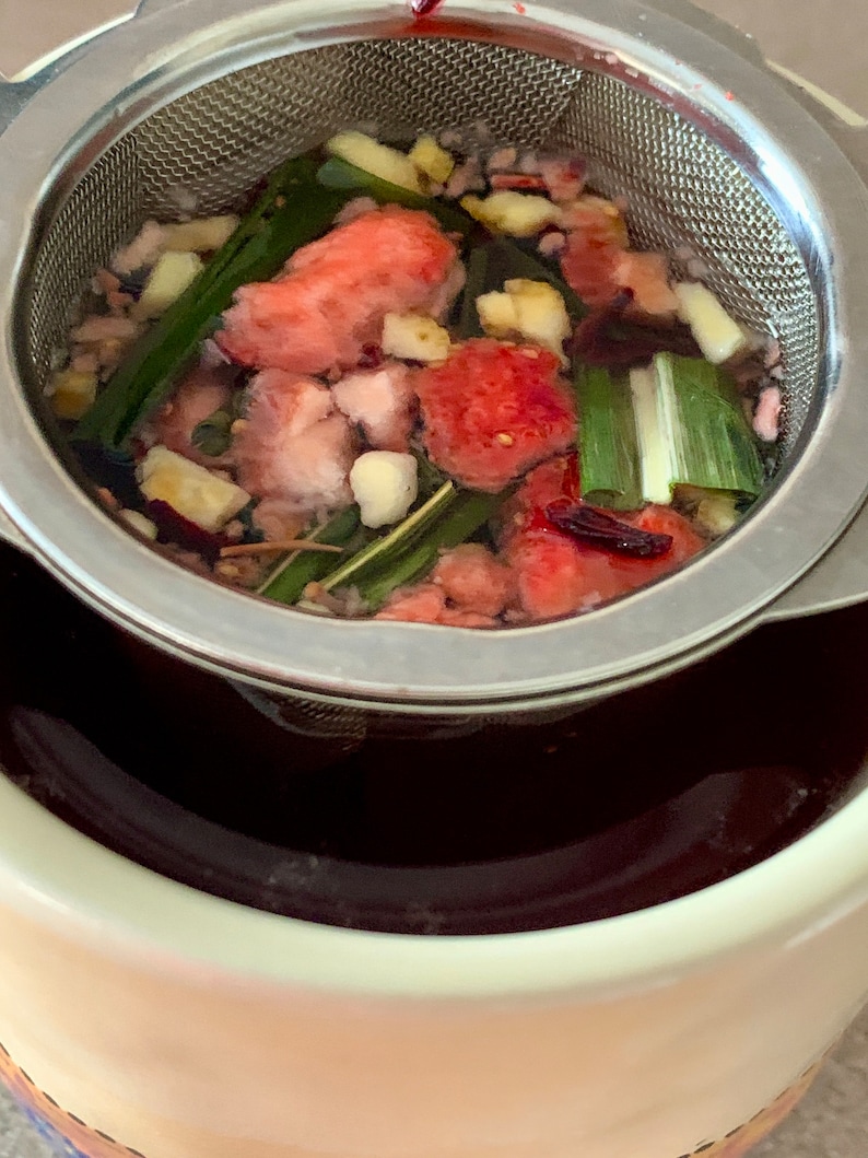 May include: A stainless steel tea infuser filled with a mixture of red and green ingredients, including fruit, herbs, and spices. The infuser is sitting in a white ceramic mug.