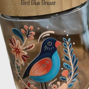 May include: A clear glass jar with a light-colored wooden lid. The jar features a colorful graphic of a bird with a blue breast, orange wing, and black head, surrounded by stylized floral and leaf illustrations. The text Bird Blue Breast is printed.