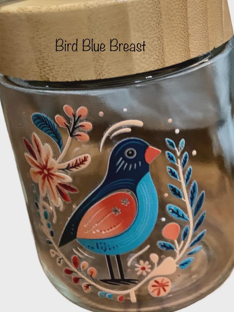 May include: A clear glass jar with a light-colored wooden lid. The jar features a colorful graphic of a bird with a blue breast, orange wing, and black head, surrounded by stylized floral and leaf illustrations. The text Bird Blue Breast is printed.