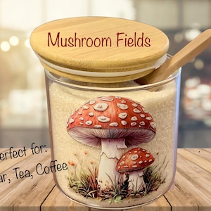 May include: A clear glass canister filled with granulated sugar, featuring a wooden lid and a small wooden spoon. The front of the canister displays a graphic of two red and white mushrooms. The text Mushroom Fields is on the lid.