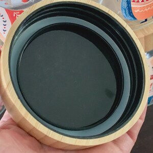 May include: A circular container lid featuring a light-colored wood grain exterior and a smooth black interior. The lid has a threaded rim designed for a secure fit, showcasing a clean, functional design with a natural wood finish and dark plastic.