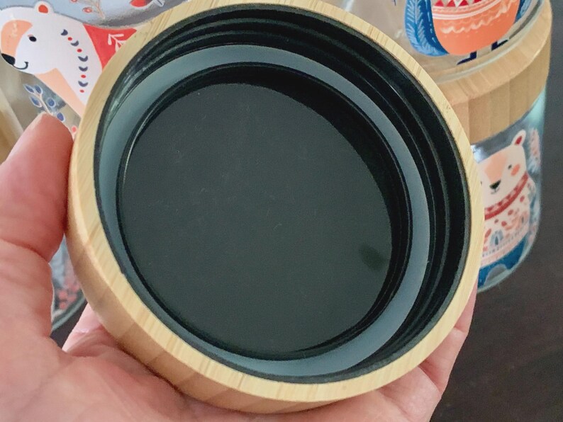 May include: A circular container lid featuring a light-colored wood grain exterior and a smooth black interior. The lid has a threaded rim designed for a secure fit, showcasing a clean, functional design with a natural wood finish and dark plastic.