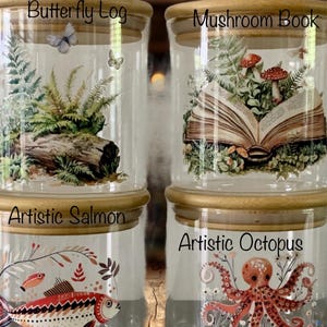 May include: Four clear glass storage jars with light wood lids. The top left jar features a log, ferns, and butterflies labeled Butterfly Log. The top right jar shows an open book with mushrooms labeled Mushroom Book. The bottom left jar displays a patterned fish labeled Artistic Salmon, and the bottom right jar shows an octopus and plants labeled Artistic Octopus.