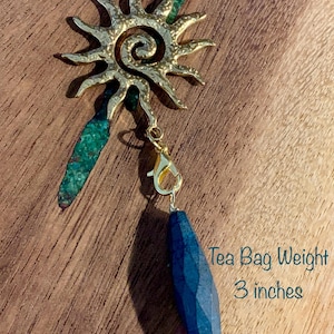 May include: A tea bag weight measuring 3 inches in total length. The item features a gold-toned sun-shaped charm with a spiral center, accented by a textured green piece and a faceted, elongated dark blue stone attached by a gold lobster clasp.