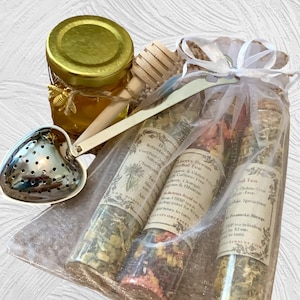 May include: A white organza bag filled with three glass tubes containing loose leaf tea blends. The tea blends are labeled "Berry Dream Herbal Tea", "Intestinal Detox Herbal Tea", and "Promote Sleep Herbal Tea". The bag also includes a honey jar, a wooden honey dipper, and a heart-shaped tea infuser.