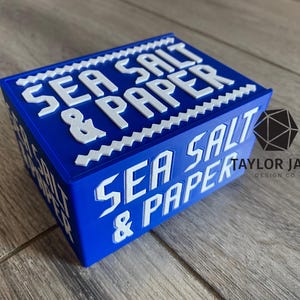 May include: A blue rectangular box with the words "SEA SALT & PAPER" in white, bold, capital letters. The box has a white zig-zag border. The words are repeated on the sides. The Taylor Jane Design Co. logo is on the side.