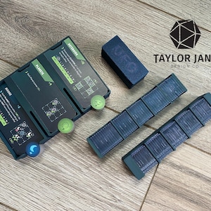 May include: A board game set with dark teal components. The set includes a card holder with game cards, two long track pieces, a small box, and two translucent green and blue game pieces. The game cards have text and graphics.