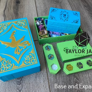 May include: A bright blue rectangular box with a gold phoenix emblem and ornate filigree, accompanied by a green storage tray containing small game pieces, two green hexagonal component holders, and a green box labelled Taylor Jane Design Co.