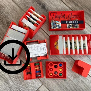 Puede incluir: Cajas de almacenamiento de plástico rojo para piezas de juego, cartas y fichas. Las cajas están etiquetadas con "Taylor Jane Design Co."