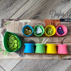 May include: A board game organizer with various colored containers holding game pieces. The containers are blue, green, yellow, and pink. The game pieces include green leaf shapes, gold gears, and blue marbles. The board has the text "Taylor Jane Design Co."