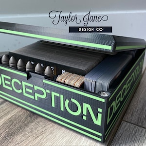 May include: A black and green custom game box with the word "DECEPTION" printed on the front. The box is open and contains a deck of cards and several small, gray, bullet-shaped objects.